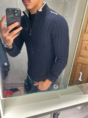 Marinblå ribbad kofta från GANT - Snygg marinblå kofta från GANT i storlek M. Tröjan är ribbstickad med vit kontrast på insidan av kragen och längs axlarna. Den har lång ärm och dragkedja framtill, perfekt för lager-på-lager. Passformen är slim och materialet känns mjukt och stickat.