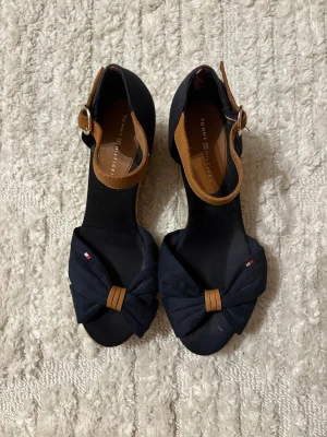 Tommy Hilfiger kilklackar amarinblå - Snygga marinblå sandaler från Tommy Hilfiger med kilklack i repdesign. Skorna har öppen tå, rosettdetalj framtill och bruna remmar i skinnimitation med spänne runt ankeln. Perfekta för sommarens alla outfits.