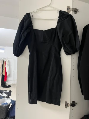 Svart kort klänning med puffärm - Snygg svart klänning med puffiga korta ärmar och fyrkantig halsringning. Modellen är kort och har en figurnära passform som ger en trendig siluett. Perfekt för dig som gillar statement-ärmar och stilren design. Materialet känns lätt och har en matt finish.