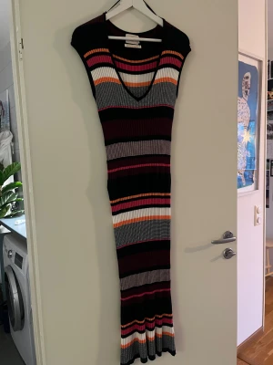 Randig långklänning Holly & Whyte - Supersnygg långklänning från Holly & Whyte med coola ränder i svart, vit, orange, rosa och grå. Klänningen är ribbad och har en väldigt fin figur då den är lös upp till och kramar om i midjan  Materialet är stickat och stretchigt, vilket gör den riktigt skön och figurnära.
