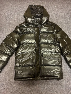 Svart pufferjacka från Prada - Säljer svart pufferjacka från Prada med glansig finish och stor huva. Jackan har två rejäla fickor framtill, dragkedja och knappar, samt ikonisk Prada-logga på bröstet. Perfekt för kalla dagar och riktigt snygg streetstil. Finns i M, L och XL. 