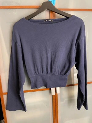 Mörkblå croppad topp Gina Tricot - Snygg mörkblå croppad topp från Gina Tricot med långa ärmar och ribbad struktur. Modellen har en bred, markerad midja och en rundad halsringning. Perfekt för dig som gillar stilrena och bekväma plagg med lite edge.