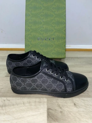 Svarta sneakers från Gucci - Snygga svarta sneakers från Gucci med klassiskt GG-mönster i grått på sidan. Skorna har rund tå, snörning och detaljer i skinn på tå och häl. Yttermaterialet är en mix av textil och skinn, vilket ger en lyxig känsla. Perfekt för dig som vill ha en stilren och exklusiv look.    Boxen tillhör inte skorna. De är helt nya använder inte de eftersom jag har andra skor som jag använder och de här skorna har varit hemma i länge. Priset är lite flexibelt 