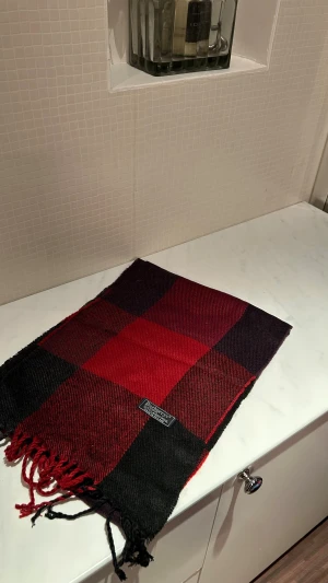 Rutig röd och svart ullväst Burberry - Snygg rutig väst från Burberry i rött och svart, tillverkad i 100% lammull. Klassisk design med stora rutor och fransar nedtill. Perfekt för dig som gillar tidlös stil med en touch av brittisk elegans.