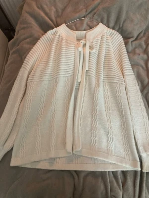 Vit stickad kofta med knytband - Superfin vit stickad kofta med snygga strukturerade ränder och vågiga detaljer. Koftan har lång ärm och ett brett ribbat parti upptill samt ett knytband vid halsen för extra stilpoäng. Den har ingen lapp var den kommer ifrån eller vilken storlek men skulle säga S/M.