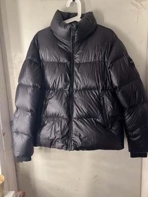 Svart pufferjacka  - Säljer en svart glansig pufferjacka från 8848 Altitude med hög krage och snyggt emblem på ärmen. Jackan har dragkedja framtill, två sidofickor med dragkedja och ribbade muddar vid ärmslut. Materialet är glansigt syntet och jackan är quiltad för extra värme. Använd 1 gång så nästan helt oanvänd!:)
