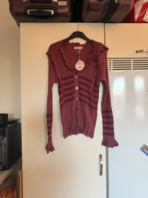 Damig kofta från Odd Molly i vinrött - Supersöt lila/vinröd kofta från Odd Molly med ribbad struktur, volangkant vid halsen och ärmslut samt knappar framtill. Koftan har långärmade ärmar med mönstrade ränder och en feminin passform. Perfekt för dig som gillar detaljer och vill sticka ut lite extra. Storlek 4 som enligt deras storleksguide motsvarar en storlek L/XL. Helt ny med tags/ prislapp fortfarande kvar. 