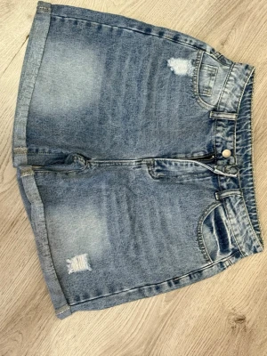 Blå denimshorts med slitningar, M - Snygga blå denimshorts i klassisk femficksmodell med slitna detaljer fram och bak. Shortsens material är 86% bomull, 6% polyester och 8% viskos. De har hög midja, dragkedja och knapp framtill samt bälteshällor. Perfekta för en chill sommardag.