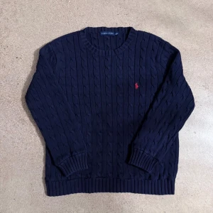Marinblå stickad tröja Ralph Lauren - Klassisk marinblå stickad tröja från Ralph Lauren med kabelstickat mönster och rund hals. Tröjan har långa ärmar och ribbade muddar vid ärmslut och nederkant. Röd broderad logga på bröstet ger en snygg kontrast. Super skick den är som ny. Passar Storlek S vuxen.