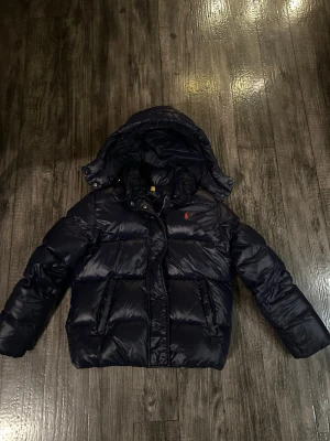 Mörkblå pufferjacka från Polo Ralph Lauren - Säljer en mörkblå pufferjacka från Polo Ralph Lauren med huva och klassisk röd logga på bröstet. Jackan har långa ärmar, två sidofickor och quiltad design. Perfekt för kalla dagar och har en snygg, glansig finish. Jackan har en minimalistisk defekt på vänster arm som inte syns ifall man inte letar efter den, samt att ”hängaren” har gått upp på ena sidan, men fixas extremt enkelt med nål och tråd💙storlek 12-14 år/ xs säljer billigt då jag vill bli av med den så snabbt som möjligt💙