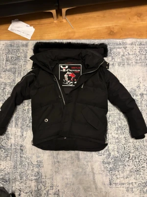 Svart Moose Knuckles dunjacka S - Svart dunjacka från Moose Knuckles med fet broderad hockeypatch på insidan. Jackan har pufferstil, dragkedja, knappstängning och fickor med lock. Huva med svart fuskpäls och coola detaljer som metallöglor och innerfickor. Perfekt för kalla dagar.