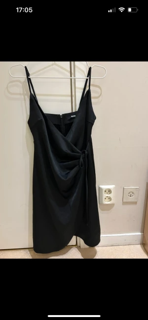  Ny Svart omlottklänning från Bik Bok - Supersnygg svart kortklänning från Bik Bok med omlottdetalj och knyt i midjan. Klänningen har tunna axelband och dragkedja bak. Materialet är mjukt och har en lätt satinliknande finish som ger en cool vibe. Perfekt för dig som gillar stilrena och trendiga plagg.