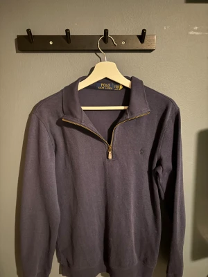 Mörkblå half zip tröja Polo Ralph Lauren - Snygg mörkblå långärmad tröja från Polo Ralph Lauren med half zip-dragkedja i guld och broderad logga på bröstet. Tröjan har en klassisk krage och är gjord i mjuk bomull, perfekt för chill dagar. Passar dig som gillar stilren och enkel design.