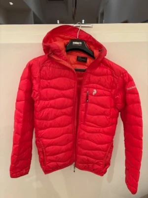 Röd dunjacka Peak Performance S - Säljer en röd dunjacka från Peak Performance i storlek S. Jackan har huva, dragkedja framtill och en bröstficka med zip. Den är quiltad och har en sportig look, perfekt för kalla dagar. Märket syns på bröstet och ärmen. R&D-logga på huvan.