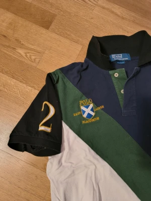 Pikétröja Polo Ralph Lauren blå/grön - Säljer en snygg pikétröja från Polo Ralph Lauren med diagonal färgblock i marinblått, grönt och vitt. Tröjan har korta svarta ärmar, broderad logga och nummer 2 på ena ärmen. Klassisk krage och knappar framtill. Perfekt för dig som gillar sportig stil.