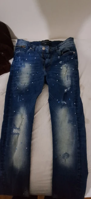 Blå jeans Supply & Demand 30W - Snygga blå jeans från Supply & Demand i storlek 30W. De har en cool tvättad look med slitningar och färgstänk på framsidan, klassiska fem fickor och knappstängning. Passformen är rak och materialet är klassisk jeansbomull.