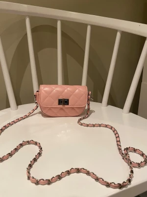 Rosa quiltad axelväska med kedja - Supersöt liten axelväska i ljusrosa med quiltat mönster och silverfärgad kedjerem med invävt rosa band. Väskan har ett snyggt spänne framtill och rundad form. Perfekt för att lyfta din outfit med en poppig färg och trendig look.
