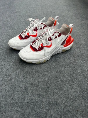 Nike React Vision sneakers vit/röd - Nike React Element sneakers i vitt med röda och vinröda detaljer. Ovandel i mesh med synliga sömmar, vita snören och en chunky sula med React-dämpning. Snygg design med kontrasterande färgblock och genomskinliga partier på sidan.
