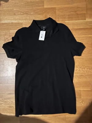 Svart piké från H&M, storlek S - Svart pikétröja från H&M i storlek S. Klassisk krage och korta ärmar, perfekt för en clean och enkel look. Regular fit och tillverkad i mjukt bomullsmaterial. Snygg att matcha med jeans eller shorts för en chill stil.