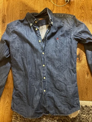 Blå skjorta från Morris Stockholm - Snygg blå skjorta från Morris Stockholm i klassisk design med knappar framtill och button-down krage. Skjortan har ett litet rött broderat emblem på bröstet och är tillverkad i mjuk bomull. Perfekt för dig som gillar stilrena och tidlösa plagg.