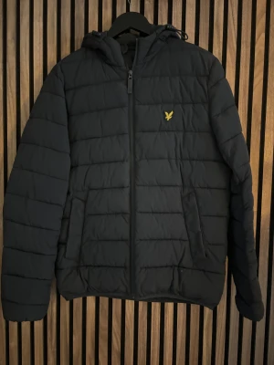 Svart pufferjacka från Lyle & Scott - Svart pufferjacka från Lyle & Scott med gul logga på bröstet. Jackan har huva, två sidofickor och dragkedja framtill. Quiltad design med horisontella sömmar och långärmad passform. Perfekt för kalla dagar och enkel att matcha med din stil. Kan sänka pris