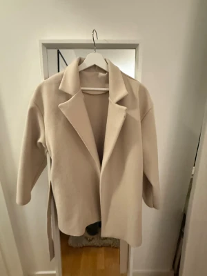 Beige kappa med bälte och krage - Snygg beige kappa med klassisk krage och bälte i midjan. Jackan har en stilren och rak passform, perfekt för dig som gillar minimalistisk stil. Materialet känns mjukt och har en ull-liknande look. Långärmad och utan synliga knappar framtill.