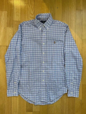 Blårutig skjorta från Ralph Lauren - Snygg blårutig skjorta från Ralph Lauren i slim fit med klassisk krage och knappar framtill. Skjortan har det ikoniska broderade Polo-logot på bröstet och är gjord i 100% bomull för skön känsla. Perfekt för dig som gillar stilrena och tidlösa plagg.