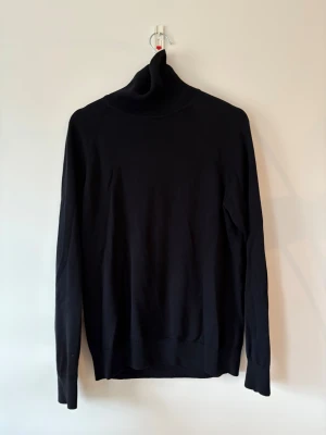 Svart polotröja från Bläck, storlek M - Svart polotröja från Bläck i storlek M. Tröjan har en hög krage och är långärmad med ribbade muddar vid hals och ärmslut. Materialet är mjukt och känns skönt mot huden, perfekt för kyliga dagar. Enkel och stilren design som funkar till många olika looks.