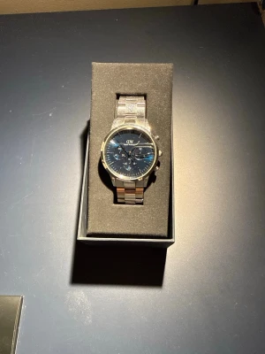 Mörkblå klocka Daniel Wellington   - Stilren herrklocka från Daniel Wellington  säljes med metallarmband och mörkblå urtavla. Tillkommer med  originalförpackning och inkluderar extra länkar, verktyg för justering samt bruksanvisning och garantiinformation. Bra skick men tecken på användning förekommer. Ny pris ca 3199 kr Hör av er vid intresse!