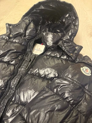 Svart pufferjacka från Moncler - Snygg svart pufferjacka från Moncler med glansig finish och klassisk logga på ärmen. Jackan har huva, dragkedja och quiltad design som håller värmen. Perfekt för kalla dagar och riktigt trendig look.