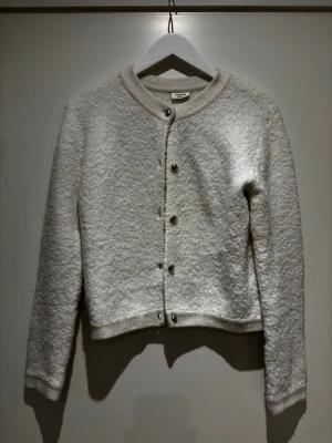 Vit bouclékofta med knappar - Säljer en stilren vit kofta i bouclématerial med rund halsringning och silvriga knappar framtill. Koftan är kortare i modellen och har långa ärmar. 