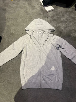 Ljusgrå stickad hoodie med dragkedja från RYVLS - Ljusgrå hoodie från RYVLS med dragkedja framtill och justerbar huva med snören. Tillverkad i mjukt material och har ribbade muddar vid ärmslut och nederkant. Perfekt för en avslappnad stil.