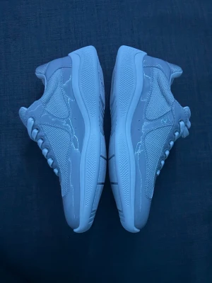 Vita sneakers från Prada med röd detalj - Snygga vita sneakers från Prada med mesh och skinnpaneler. Skorna har en röd detalj på hälen och silvriga partier upptill. Klassisk låg modell med snörning och rund tå. Perfekta för dig som gillar stilrena och exklusiva sneakers.