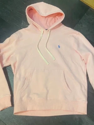 Ljusrosa hoodie från Polo Ralph Lauren - Ljusrosa hoodie från Polo Ralph Lauren med klassisk huva och dragsnören. Broderad blå logga på bröstet och stor känguruficka framtill. Mjuk bomullskvalitet och ribbade muddar vid ärmslut och nederkant.