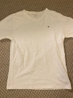 Vit v-ringad t-shirt Tommy Hilfiger - Klassisk vit t-shirt från Tommy Hilfiger med v-ringning och liten broderad logga på bröstet. Tillverkad i mjuk bomull och har en enkel, stilren design som passar till allt. Storlek 12-14år