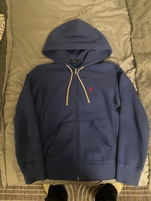Blå hoodie från Polo Ralph Lauren - Blå zip-hoodie från Polo Ralph Lauren med vit dragsko och klassisk röd logga på bröstet. Mjuk bomullskänsla, känguruficka och ribbade muddar. Perfekt för en chill och stilren look.