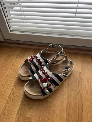 Inuikii Marinblå Beige Sandaler - Inuikii tofflor sandaler i marinblå beige / cream / vit med röd och vit juveler och guld detaljer Spännena kan användas för att dra åt eller lossa skon beroende på önskad passform. Storlek 40 använt för en sommar köpte tyvärr fel storlek. Lite vatten fläckar i en sko och vatten märke i den andra. Syns på bilderna påverka ej användning. Kan skicka fler bilder vid behöv. 