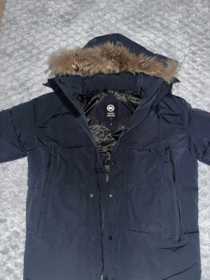 Mörkblå parkas från Canada Goose - Snygg mörkblå parkas från Canada Goose med stor huva och avtagbar fuskpäls.  Det är fel på storlekn det står S men den är mycket större en vad som står 