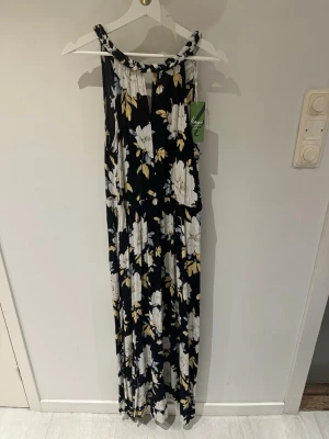 Blommig långklänning från Vila storlek 38 - Snygg långklänning från Vila med svart botten och stora vita blommor med inslag av beige och gul. Klänningen har ärmlös design och rund halsringning. Perfekt för sommarens alla tillfällen och tillverkad i ett lätt, luftigt material.
