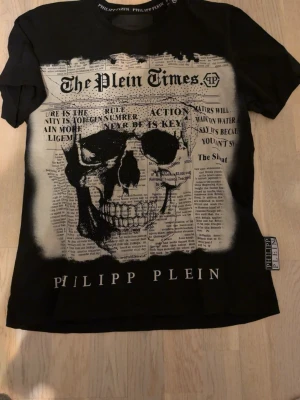 Svart Philipp Plein t-shirt med döskalle - Cool svart t-shirt från Philipp Plein med stort tryck av en döskalle och tidningsinspirerad text på framsidan. Rund halsringning med logga på insidan och en extra etikett på sidan. Perfekt för dig som gillar edgy streetwear. Äkta ! Pris går o diskutera vid snabb affär!