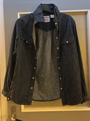 Mörkblå jeansskjorta - Klassisk mörkblå jeansskjorta med tryckknappar och två bröstfickor. Skjortan har lång ärm och en avslappnad passform, perfekt till jeans eller över en t-shirt.