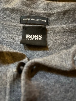 Grå stickad tröja från Hugo Boss - Snygg grå stickad tröja från Hugo Boss i mjukt italienskt garn. Tröjan har krage och två knappar vid halsen för en stilren look. Ribbstickade muddar vid ärmslut och nederkant. Perfekt för dig som gillar klassisk och enkel stil.