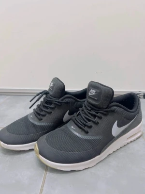 Nike Air Max Thea svarta sneakers - Nike Air Max Thea sneakers i svart med vit sula och klassisk Nike-logga på sidan. Skorna har snörning och en låg profil med rund tå. Ovandelen är i syntetmaterial och mesh för bra andningsförmåga. Perfekta för dig som gillar stilrena och sportiga sneakers.