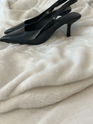 Svarta lackade pumps med klack - Säljer ett par svarta pumps i lackat material med spetsig tå och öppen häl. Skorna har en elegant smal klack och stilren design som passar perfekt till alla outfits. Klassisk och trendig modell för dig som gillar att sticka ut.