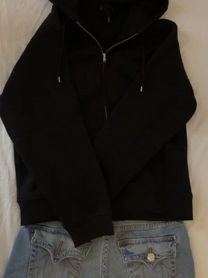 Svart hoodie med dragkedja från H&M - Klassisk svart hoodie från H&M med dragkedja framtill och justerbar huva. Tillverkad i mjukt bomullsmaterial och har ribbade muddar vid ärmslut och nederkant. Perfekt för en chill och avslappnad stil.