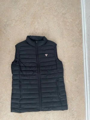 Svart dunväst från Guess - Snygg svart dunväst från Guess med quiltad design och diskret logga på bröstet. Västen har dragkedja framtill och två praktiska fickor med dragkedja. Perfekt för lager på lager och en clean streetstyle-look.