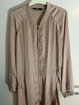 Beige bomberjacka klännning från Bik Bok - Stilren beige bomberjacka klänning från Bik Bok med lång ärm, ribbade muddar och dragkedja framtill. Jackan har en lätt glansig yta och två fickor på sidorna. Perfekt för dig som gillar en enkel men trendig look.