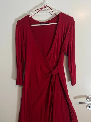 Röd omlottklänning med knyt i midjan från Indiska - Snygg röd omlottklänning från Indiska med 3/4-ärmar och djup V-ringning. Klänningen har ett knyt i midjan som ger en smickrande siluett och är gjord i ett mjukt, stretchigt material som känns bekvämt mot huden.