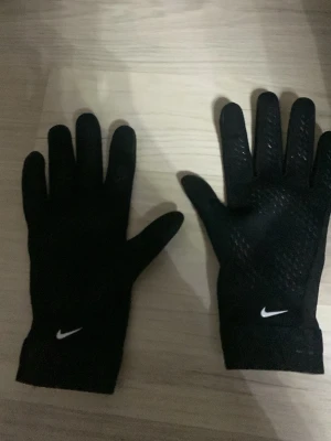 Svarta Nike löparhandskar - Svarta sporthandskar från Nike med vitt logotryck och mönstrade detaljer på handflatan och fingrarna för extra grepp. Perfekta för löpning eller träning utomhus. Snygg och smidig design som passar till sportiga outfits.