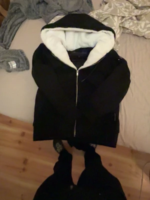 Svart vindjacka med vit huva - Snygg svart vindjacka med stor huva och vit, fluffig insida. Jackan har dragkedja framtill och en relaxed passform. Perfekt för kyliga dagar när du vill hålla stilen enkel och clean.